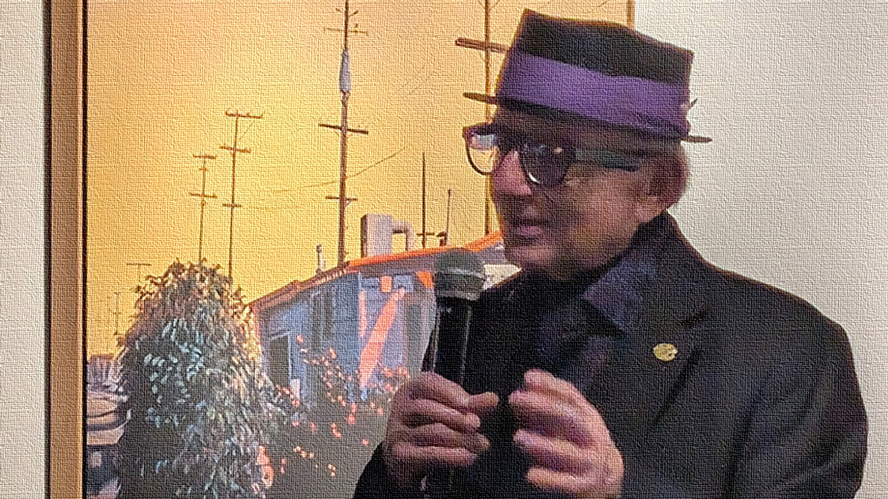 A photograph of Richard Perri with a microphone in front of one of his yellow-heavy paintings. Perri is an elderly gentleman with greying hair who wears black, thick-rimmed glass; a black hat with a royal purple band; a dark grey suit; a black button-up with royal purple flowers; and a gold pin on his lapel. The photograph has been edited to look like it is on canvas.