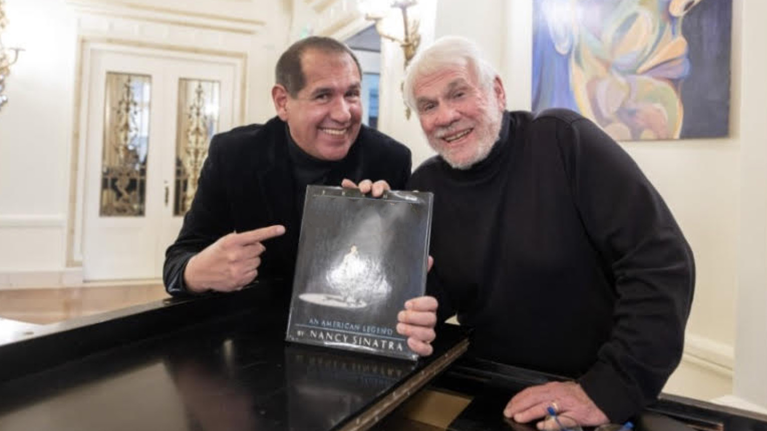 A photograph of two men standing at a piano and holding a book on top of it. The book is "Frank Sinatra: An American Legend" by Nancy Sinatra and has an image of Frank singing in the spotlight. The man to the left points at the book. He wears an all-black suit and smiles; his short hair is brown. To the right is an elderly man wearing a black turtleneck. He has white hair and a white beard.