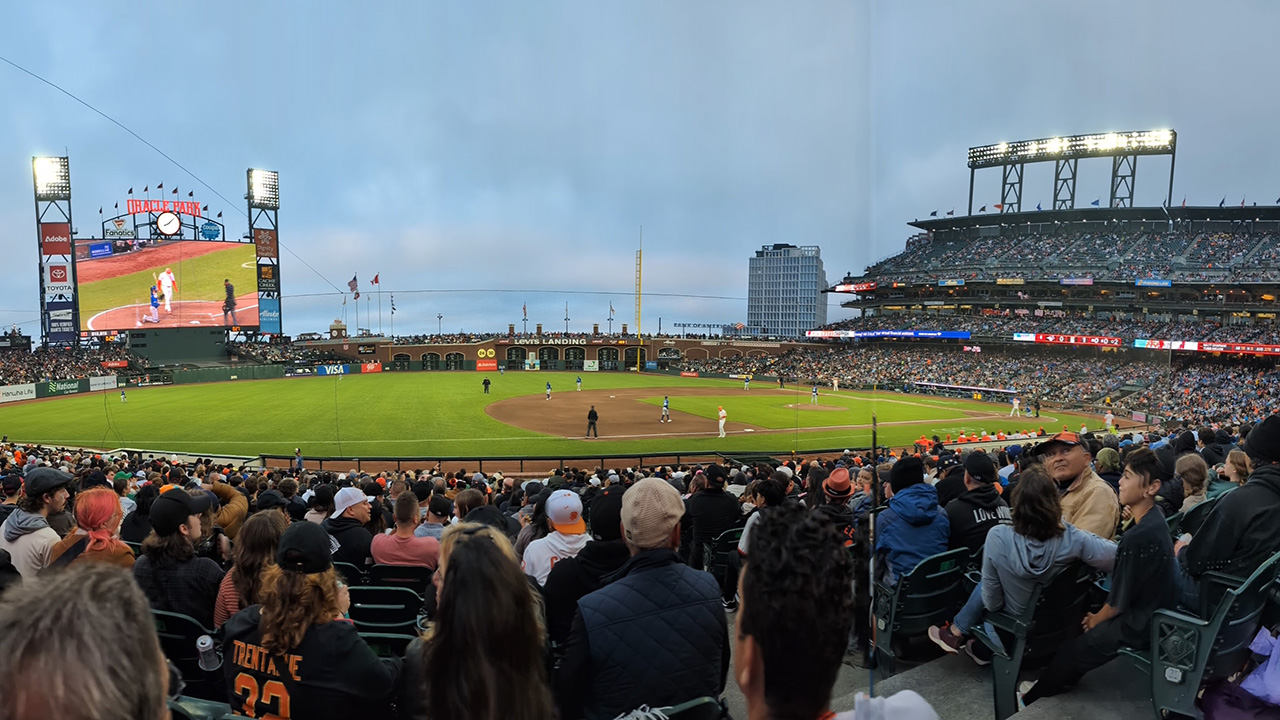 A photo from the bleachers of Oracle Park during a Giants game. The filled stands watch the baseball field below, as well as the stadium's big screen, as a man with a bat gets ready for the pitcher to throw a baseball.