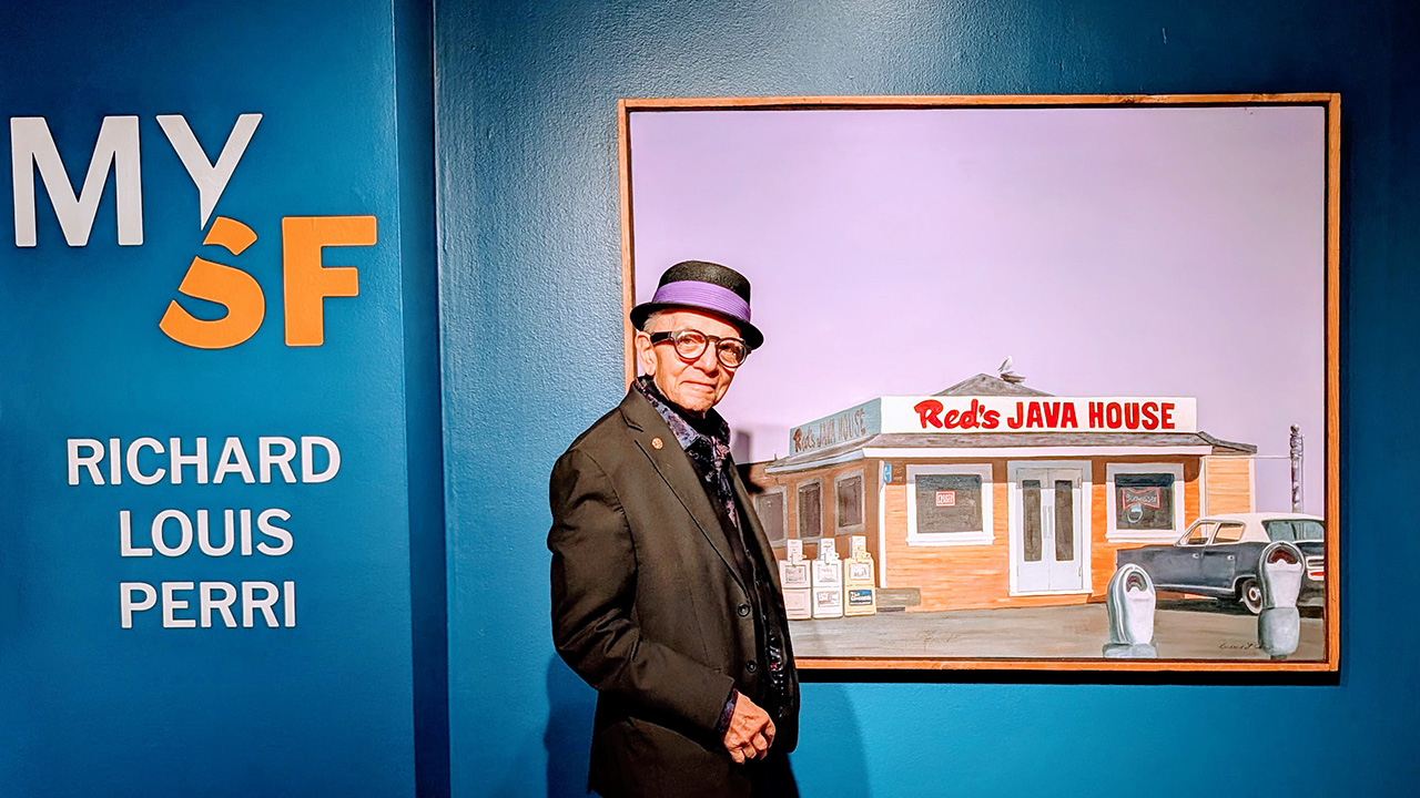 Richard Perri stands in front of the title wall of his exhibition "My SF" next to his painting of Red's Java House. Perri is an elderly gentleman with greying hair who wears black, thick-rimmed glass; a black hat with a royal purple band; a dark grey suit; a black button-up with royal purple flowers; and a gold pin on his lapel. His painting shows Red's Java House, a brown building, below a lavender sky. An old blue & white Chevrolet is parked in front of it. The wall the painting hangs on is a dark blue.