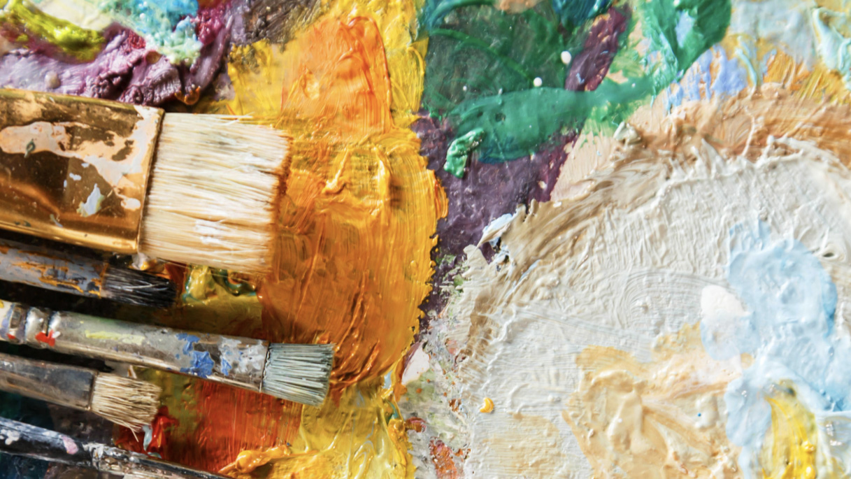 A close-up of an oil paint palette with dried paints on it. The most prominent colors are white, brown, orange, yellow, purple, and teal. Emerging from the lower left side of the image are five paint brushes with paint on the metal and wood parts of the brush. The bottom four brushes are of a similar size, but the top brush is large and flat.