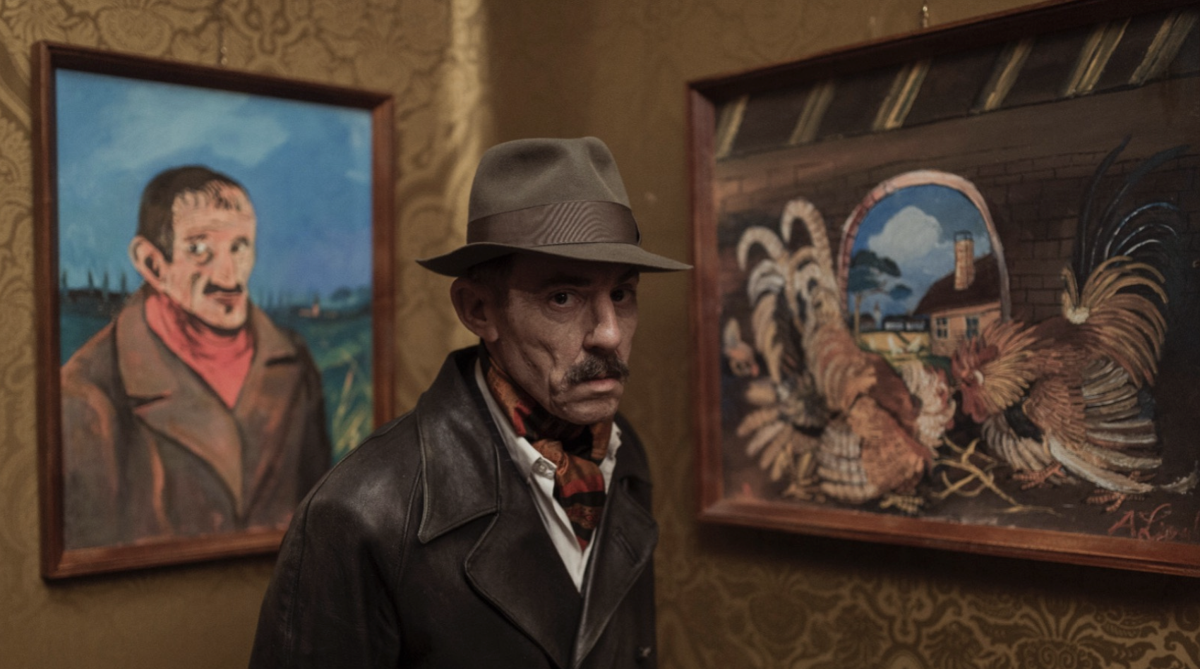 An elderly man, Elio Germano playing Antonio Ligabue, stands in front of the corner of a golden patterned wall. Shown only from the chest up, he wears a dark brown leather jacket; a white button-down shirt slightly open at the collar; a red, orange, and black patterned scarf tucked into the open collar; and a brown fabric fedora. The 3/4 bust faces the right of the image, and his brown eyes stare at the viewer. A full, brown mustache sits above his frown, his hat casting a severe shadow across his thin face. To his left is a framed painting, presumably a self-portrait of himself in similar attire, without a hat to cover his balding brown hair, against a background of green farmland and blue sky. To his right is a framed painting of a hen and a rooster with fluffed feathers in front of a small portal window that has a view of a cloudy blue sky, and orange house with a chimney, a yellow wall, and a tree.