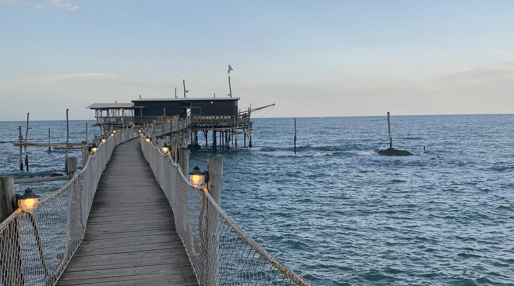 A photograph of a thin, wooden walkway with netted railing that leads to a small pier with a brown building. Small lights along the walkway suggest it is almost dusk. The blue ocean surrounds the pier and walkway on all sides. Waves crash against a rock to the right of the pier. Above the horizon line is a blue sky with one or two white clouds.