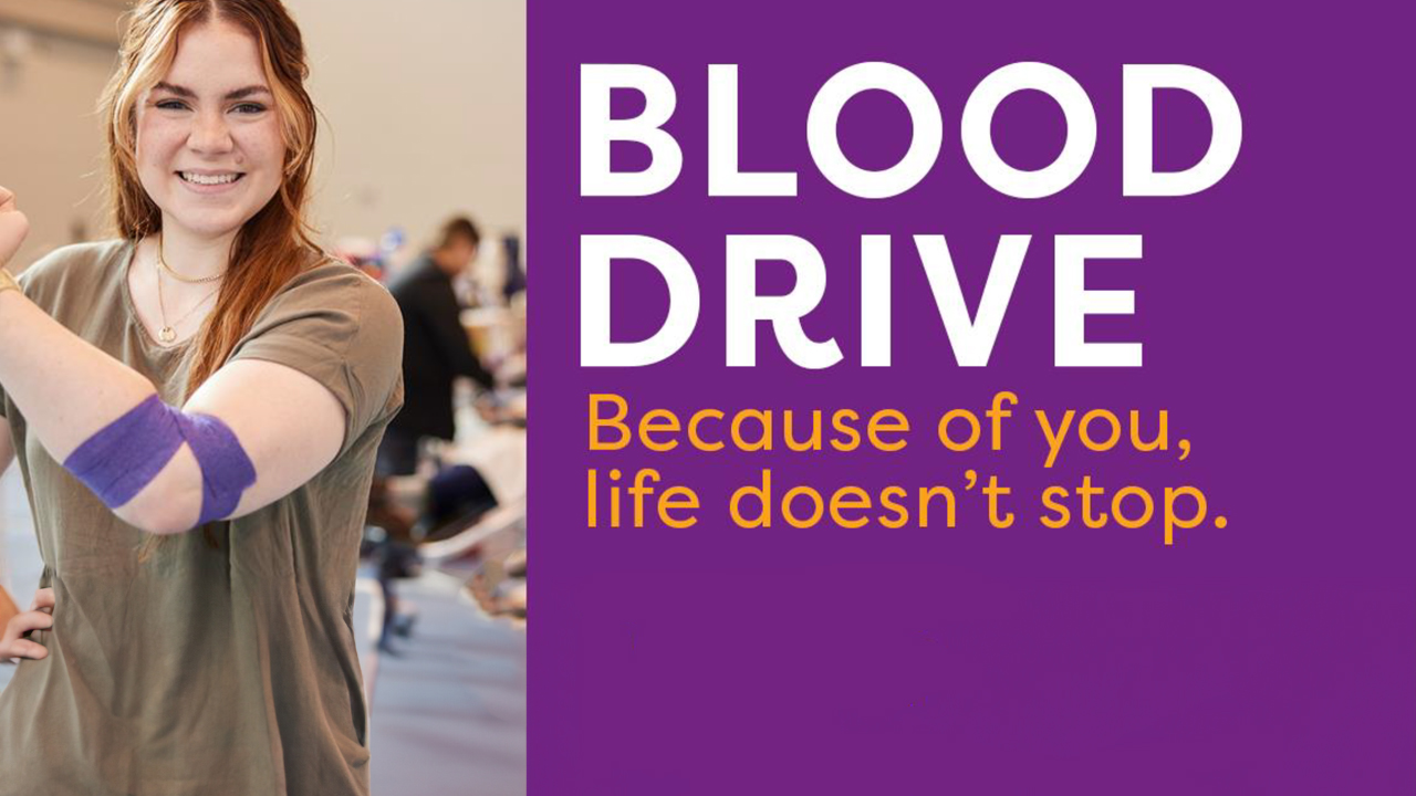Bold, white lettering reads "BLOOD DRIVE" against a royal purple background. Golden yellow text directly below states "Because of you, life doesn't stop." To the left of the text is an image of a grinning woman with red hair and a greyish-green t-shirt standing and flexing one arm directly in front of her torso. She has purple medical tape wrapped around her elbow. Her other hand rests on her hip. In the blurry background, people get their blood drawn.